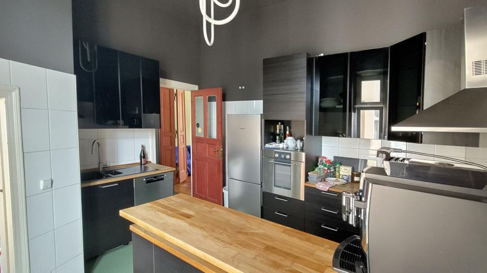 Moderne Küche mit schwarzen Hochglanzschränken, weißer Fliesenrückwand und zentraler Holzarbeitsinsel. Sichtbar sind Herd mit Edelstahl-Dunstabzug, Kühlschrank und Spüle; eine rote Innentür führt in den angrenzenden Bereich.