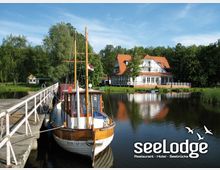 Ein weißes Fachwerkgebäude mit rotem Dach, umgeben von Bäumen, liegt am Ufer eines Sees. Im Vordergrund ein hölzernes Boot, das an einem Steg angelegt ist, mit ruhigem Wasser und blauem Himmel im Hintergrund.