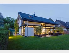 Modernes Einfamilienhaus mit Satteldach und großer Glasfront, beleuchtet in der Dämmerung. Im Vordergrund liegen ein gepflegter Rasen und eine Terrasse mit Outdoor-Möbeln, umgeben von Hecken und Nachbarhäusern.