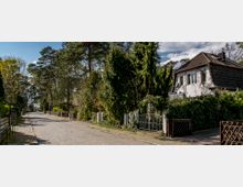 Einfamilienhaus mit weißer Fassade und dunklem Dach in einer ruhigen Wohnstraße mit Kopfsteinpflaster, umgeben von Bäumen, hohen Hecken und Gartenzäunen, unter einem teils bewölkten Himmel.