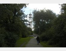 Ein Fußweg führt durch eine von Bäumen und Sträuchern gesäumte Grünanlage zu einem modernen Kirchturm aus Beton mit einem Kreuz an der Spitze. Im Vordergrund gehen zwei Personen auf dem Weg in Richtung des Turms.