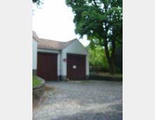 Zwei kleine Gebäude mit weißen Wänden und braunen Doppeltüren, gelegen an einem gepflasterten Hof. Im Hintergrund sind Bäume und grüne Vegetation sichtbar.