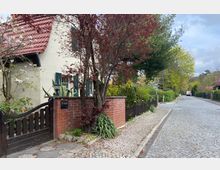 Straßenszene in einem Wohngebiet mit gepflastertem Gehweg und Straße; links ein Haus aus hellen Steinen mit roten Dachziegeln und grünen Fensterläden, umgeben von einer roten Backsteinmauer und Holzzaun. Im Vordergrund stehen Bäume und Pflanzen, im Hintergrund sind weitere Häuser und Grünflächen zu erkennen.