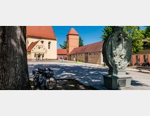 Großer gepflasterter Platz mit historischen Gebäuden mit roten Ziegeldächern, darunter eine Kirche mit Rundbogenportal und ein turmartiger Bau. Im Vordergrund stehen ein steinernes Denkmal mit Relief eines Adlers sowie Fahrräder neben einem großen Baum; im Hintergrund sind Bäume und weitere Gebäude zu sehen.