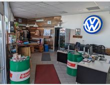 Ein Verkaufsraum mit Volkswagen-Logo an der Wand, ausgestattet mit Holzelementen, zwei Schreibtischen mit Computern, und Dekorationen wie Ölfässern und Regalen mit Produkten. Links befindet sich eine Eingangstür mit Glasfront, und im Hintergrund sind Sitzgelegenheiten sowie ein kleiner Verkaufs- und Ausstellungsbereich zu sehen.