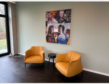 Zwei gelbe Sessel und ein schwarzer runder Beistelltisch stehen auf einem Holzboden vor einer mintgrünen Wand. An der Wand hängt ein farbenfrohes, abstraktes Gemälde, links befindet sich ein großes Fenster mit Blick ins Freie.