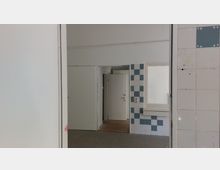 Blick in einen leeren Innenraum mit gefliesten Wänden in Weiß und Blau. Im Hintergrund befinden sich eine Tür mit Klinke, ein kleines Fenster und ein Bereich mit Holzfußboden.