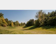 Grüne Parklandschaft mit leicht abschüssiger Wiese, umgeben von dichtem Baumbestand. Der Himmel ist klar und blau, und die Vegetation zeigt Anzeichen von herbstlicher Verfärbung.