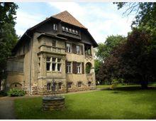Historisches zweistöckiges Gebäude mit Putzfassade und roten Ziegeldach, umgeben von einem gepflegten Garten mit Rasenfläche und hohen Bäumen. Im Vordergrund steht ein steinerner Brunnen auf einer grünen Wiese.