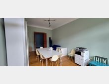 Helles Besprechungszimmer mit langen weißen Tisch und mehreren Stühlen auf Holzboden, mintgrünen Wänden und hoher Decke mit moderner Deckenleuchte. Rechts steht ein großer Kopierer neben einem Heizkörper, hinten eine Holztür und einige Büromöbel.