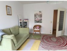 Wohnzimmer mit einem hellgrünen Sofa, einem kleinen Rattanstuhl und hellem Teppich mit roten Kreismustern. An der weißen Wand hängen dekorative Bilder, und vor dem Sofa steht ein kleiner Spiegel neben einem schmalen Regal.