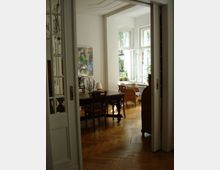 Wohnraum mit Holzparkettboden, einem schweren Holztisch mit Stühlen und einem Sessel vor großen, geöffneten Fenstern. Die weiß gestrichenen Wände und hohe Decken lassen viel Tageslicht in den Raum, der an ein Wohnzimmer oder ein Esszimmer erinnert.