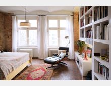 Schlafzimmer mit Backsteinwänden, großen Fenstern mit weißen Vorhängen und einem Ledersessel mit Fußablage. Ein Bücherregal und ein Schreibtisch mit Dekorationen sind ebenfalls zu sehen, zusammen mit einem bunten Teppich auf dem Boden.