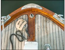 Ansicht der Spitze eines Holzboots mit lackierten Rändern und verlegtem Dielenboden. Sichtbare Details sind eine Seilhalterung, eine Metallklampe, ein zusammengerolltes Kabel sowie ein runder Gegenstand mit der Aufschrift „Marchal“.