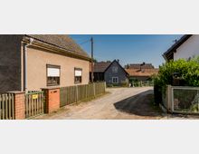 Dorfstraße mit niedrigen Wohnhäusern und Zäunen; links ein Haus mit hellbrauner Fassade und geschlossenem Rolladen, im Hintergrund weitere Häuser mit Schrägdächern. Eine unbefestigte Straße führt durch die Szene, rechts ist ein Garten mit Hecken zu sehen.