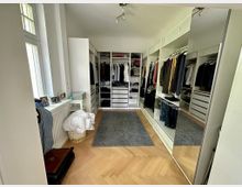 Ein begehbarer Kleiderschrank mit hölzernem Fischgrätboden, offenen Regalen, Kleiderstangen und Schubladen an beiden Seiten. Auf der rechten Seite befinden sich spiegelnde Schranktüren, und links ist ein Fenster mit einer Fensterbank, auf der persönliche Gegenstände stehen.