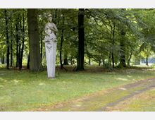 Steinfigur einer Frau auf einem hohen Sockel in einem Waldgebiet mit dichtem Baumbestand und einer grasbewachsenen Lichtung; ein schmaler Weg führt rechts an der Statue vorbei.