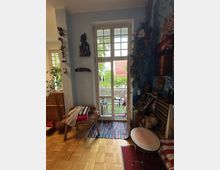 Wohnzimmer mit heller Glastür, die zu einem kleinen Garten führt. Im Raum stehen ein Holzstuhl, ein runder Beistelltisch sowie Regale mit Deko und Figuren; Wände sind mit verschiedenen Kunstobjekten und Pflanzen dekoriert. Ein bunter Teppich liegt vor der Tür auf Parkettboden.