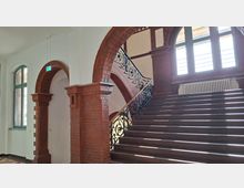 Innenansicht eines historischen Treppenhauses mit breiter Holztreppe, kunstvollem schmiedeeisernem Geländer und roten Backsteinbögen. Große Rundbogenfenster lassen viel Licht einfallen, darunter steht ein Heizkörper an der Backsteinwand.