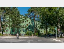 Ecke einer Wohnstraße mit einem mehrstöckigen, grün gestrichenen Wohnhaus, umgeben von hohen Bäumen. Im Vordergrund führt ein Fußgängerüberweg über eine Straßenkreuzung mit Ampeln.