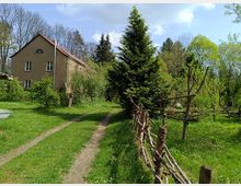 Ein ländliches Grundstück mit einem zweistöckigen, beige verputzten Haus mit rotem Satteldach im Hintergrund. Im Vordergrund verlaufen ein grasbewachsener Weg, ein niedriger Zaun aus Naturholz und verschiedene Bäume und Sträucher in einer grünen, bewachsenen Umgebung.