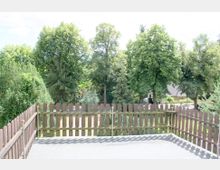 Holzzaun auf einer Terrasse mit Blick auf eine grüne Gartenlandschaft, umgeben von großen Bäumen. Im Hintergrund sind Wohnhäuser mit sichtbaren Dächern und Gehwege erkennbar.