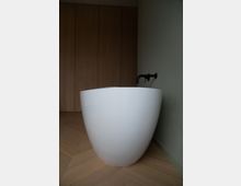 Weiße freistehende Badewanne in einem Raum mit hellen Holzböden, einer holzverkleideten Wand und einer schwarzen Wandarmatur.