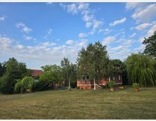 Ein einstöckiges rotes Haus mit weißen Fensterrahmen, umgeben von Bäumen, Sträuchern und Rasen. Der Himmel ist klar mit wenigen Wolken, und einige Pflanzgefäße stehen im Vordergrund.