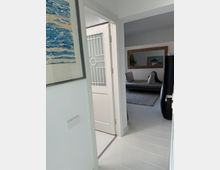 Blick in einen hellen Innenraum mit einem weißen Türrahmen, einer halboffenen Tür mit geometrischem Muster auf der Verglasung und einem angrenzenden Wohnzimmer mit Sofa, Teppich und Spiegel an der Wand. Links an der Wand hängt ein gerahmtes Kunstwerk mit blauen und beigen Mustern.