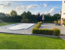 Große, gepflasterte Terrasse in einem Garten mit rechteckigem, leerem Einbaupool und einem Sonnenschirm auf der Steinfläche. Umgeben von Rasen, niedrigen Hecken, Pflanzkübeln und einer hohen Baum- und Sichtschutzhecke am Grundstücksrand.