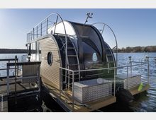 Modernes Hausboot mit geschwungenem Dach und Holzvertäfelung, festgemacht an einem Steg auf einem See. Das Boot verfügt über runde Fenster, ein Podest mit Metallgeländer sowie eine klare Sicht auf das Wasser und umliegende Uferlandschaft.