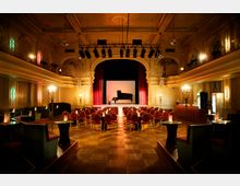 Ein historischer Konzertsaal mit red- und goldfarbenen Elementen, einer Bühne mit einem schwarzen Flügel und roten Vorhängen. Der Raum ist mit eleganten Kronleuchtern, roten Polsterstühlen und kleinen Tischen mit Kerzen dekoriert.