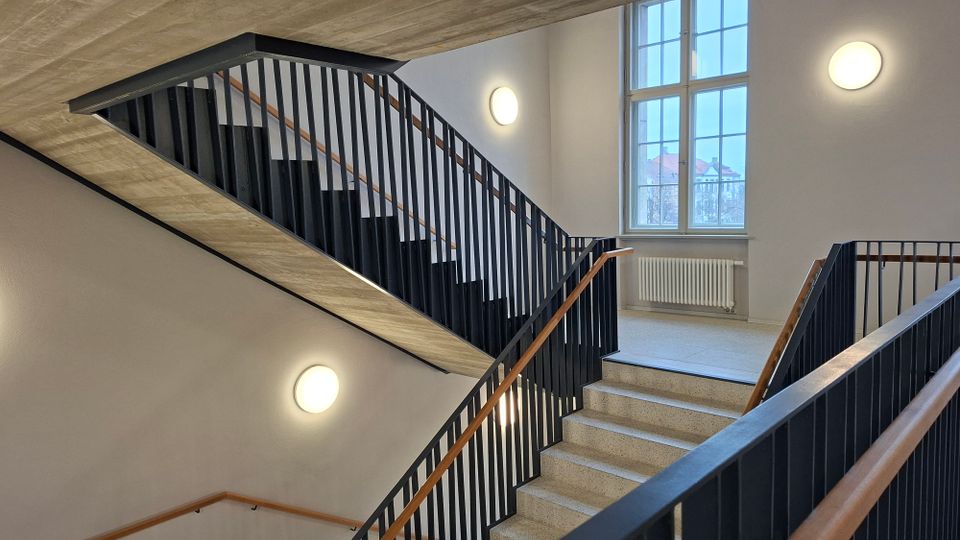 Innenansicht eines Treppenhauses mit mehreren Betonläufen, schwarzen Metallgeländern und Holzhandläufen. Helle Wände mit runden Wandleuchten sowie ein großes Sprossenfenster mit Heizkörper darunter prägen den Raum.