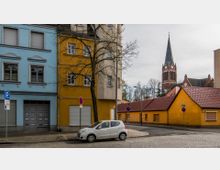 Straßenszene mit mehrstöckigen Wohnhäusern in Blau und Gelb, ein geparkter Kleinwagen auf einer gepflasterten Straße und einer gelben Gebäudereihe mit roten Dächern im Hintergrund. Eine Kirche mit rotem Backstein und spitzem Glockenturm erhebt sich dahinter, umgeben von kahlen Bäumen.