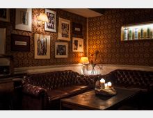 Gemütlicher Raum im Vintage-Stil mit braunen Ledersofas, einem dunklen Holztisch mit brennenden Kerzen und einer von Tapete mit einem goldenen Ornamentmuster bedeckten Wand. An der Wand hängen Bilder und ein kleines Regal mit beleuchteten Flaschen; warme Lampen sorgen für eine stimmungsvolle Beleuchtung.