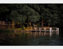 Holzsteg an einem Ufer mit Bäumen im Hintergrund, beleuchtet von warmem Licht. Im Vordergrund Wasser, am Ufer befindet sich ein Gebäude aus Backstein und Holz sowie Wimpelketten, die zwischen den Bäumen gespannt sind.