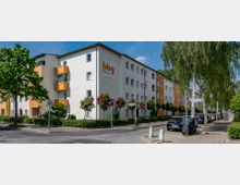 Wohngebäude in einem Wohngebiet mit weißer Fassade und orangefarbenen Balkonen. Die Straße ist gesäumt von geparkten Autos, Bäumen und Büschen; ein Straßenschild weist auf die Zobeltitzstraße hin.