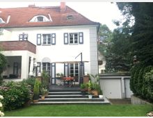 Zweistöckiges Wohnhaus mit weißer Fassade, blau gerahmten Fenstern und rotem Ziegeldach, umgeben von einem gepflegten Garten mit Rasen und Topfpflanzen. Eine Terrasse mit einer kleinen Sitzecke und Sonnenschirm führt über Stufen zum Garten, rechts daneben befindet sich eine Garage.