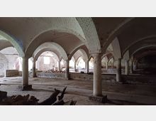 Ein historischer Gewölbekeller mit weißen Ziegelbögen, die auf dicken Steinsäulen ruhen. Der Raum ist teilweise verfallen, mit verstreuten Trümmern und schwachem Licht, das durch kleine Fenster und eine geöffnete Tür fällt.