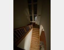 Holztreppe in einem dunklen Innenraum mit hohen Fenstern, die mit Sprossen versehen sind. Ein Heizkörper steht unter den Fenstern, und eine Wandöffnung führt in einen weiteren Raum mit rotem Bodenbelag.