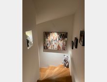 Treppenabsatz in einem modernen Innenraum mit Holzstufen, weißen Wänden und indirektem Licht. An der Wand hängt ein abstraktes Gemälde, und auf dem Boden steht eine kleine bärenförmige Figur mit schwarz-weißer Musterung.