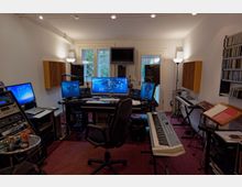 Tonstudio mit mehreren Monitoren auf einem zentralen Schreibtisch, umgeben von Lautsprechern, digitalen Audiogeräten und Aufnahmeausrüstung. Im Raum befinden sich ebenfalls ein E-Piano, ein Keyboard, Regale mit CDs und ein Notenständer; Tageslicht fällt durch eine Fenster- und Türfront mit Blick auf einen Garten.