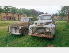 Zwei rostige, alte Pickup-Trucks stehen auf einer Wiese vor einem verfallenen, historischen Gebäude aus Backstein. Im Hintergrund sind Bäume und weitere Teile der Gebäudestruktur zu sehen.