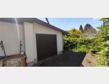 Eine kleine Garage mit braunem Tor und flachem Dach, daneben ein gepflasterter Hof, umgeben von dichtem Grün und Sträuchern. Im Vordergrund ist ein Wäschespinnenständer zu sehen.