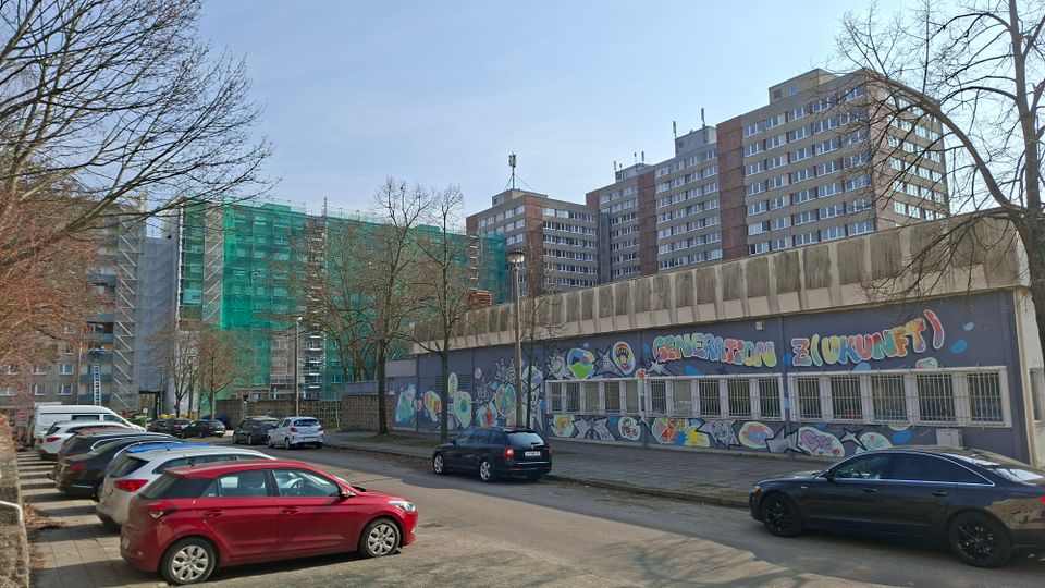 Straßenszene mit parkenden Autos vor einem langgestreckten Flachbau, dessen Fassade mit großflächigem Graffiti und vergitterten Fenstern gestaltet ist. Dahinter stehen mehrere hohe Wohn- oder Bürohochhäuser, teils mit grünem Baugerüst und Netzverkleidung, umgeben von kahlen Bäumen.