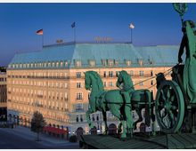 Ansicht des Hotels Adlon in Berlin bei Sonnenuntergang, im Vordergrund die Quadriga der Brandenburger Tors mit vier Pferden und der Wagenlenkerin. Das Hotel hat eine helle Fassade mit zahlreichen Fenstern und ein grünes Mansarddach, über dem National- und Europaflaggen wehen.