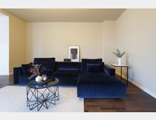 Modernes Wohnzimmer mit dunkelblauem Samtsofa, einem schwarzen Glastisch mit geometrischem Gestell und hellbeigem Teppich. Im Hintergrund eine helle Wand mit schlichter Wanddekoration und ein Beistelltisch mit Vase und Eukalyptuszweigen.