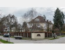Straßenszene in einem Wohngebiet mit einer großen Villa im klassischen Stil, umgeben von einer Mauer mit Buchenhecke und Zaun. Im Vordergrund steht eine Straßenlaterne und an der gepflasterten Straße sind ein geparktes Auto sowie ein Anhänger zu sehen; im Hintergrund mehrere Bäume.