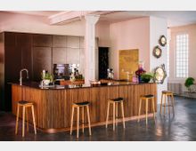 Moderne Küche mit einer großen, abgerundeten Kücheninsel aus Holz und einer dunkel glänzenden Arbeitsplatte. Im Hintergrund befinden sich Einbauschränke, ein integrierter Backofen, dekorative Wandkunst und hohe Fenster, die natürliches Licht hereinlassen.