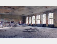 Großer, leerer, heruntergekommener Raum mit mehreren hohen Fenstern, die Tageslicht hereinlassen. Die Wände sind stark beschädigt, teilweise mit Graffiti bedeckt, und der Boden ist mit Schutt und Staub bedeckt.